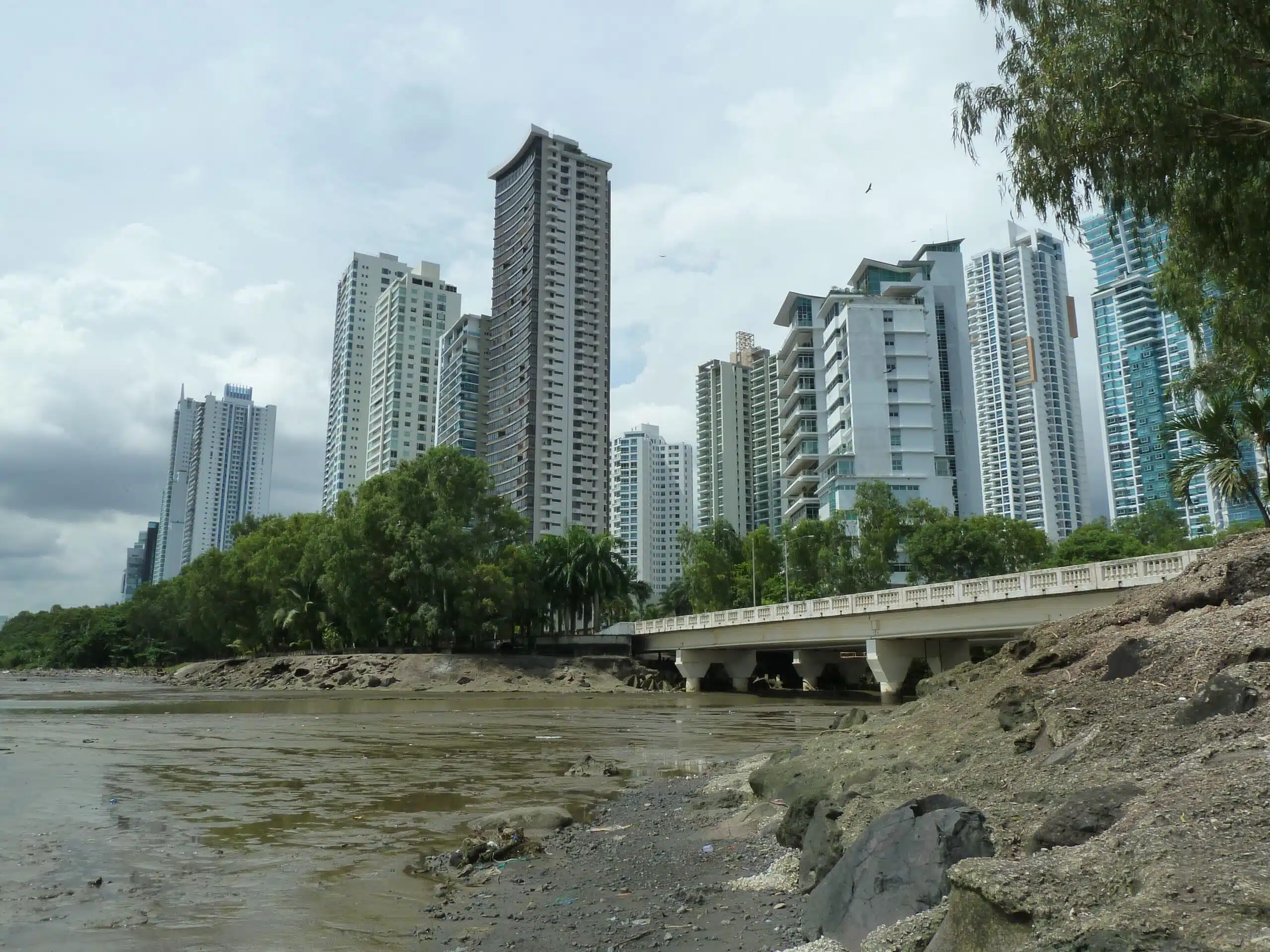 High-rise buildings along the Panama City waterfront, with green coastal vegetation beside a bridge and an exposed muddy shoreline at low tide under a cloudy sky.