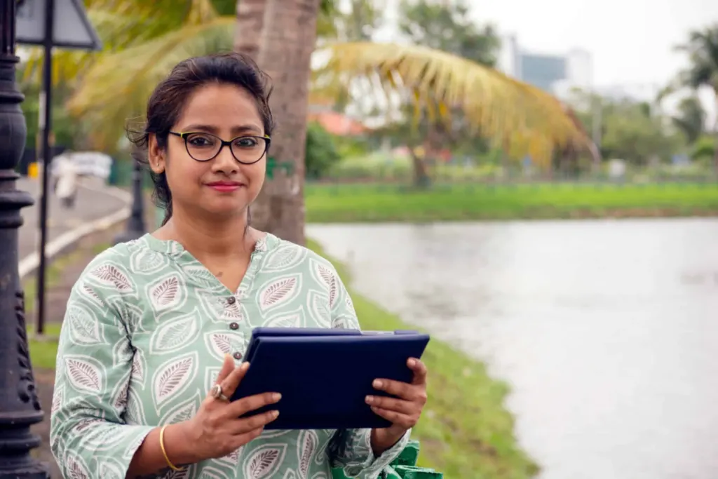 Woman looking into the camera, holding a tablet computer. With palm trees and a water body in the background.