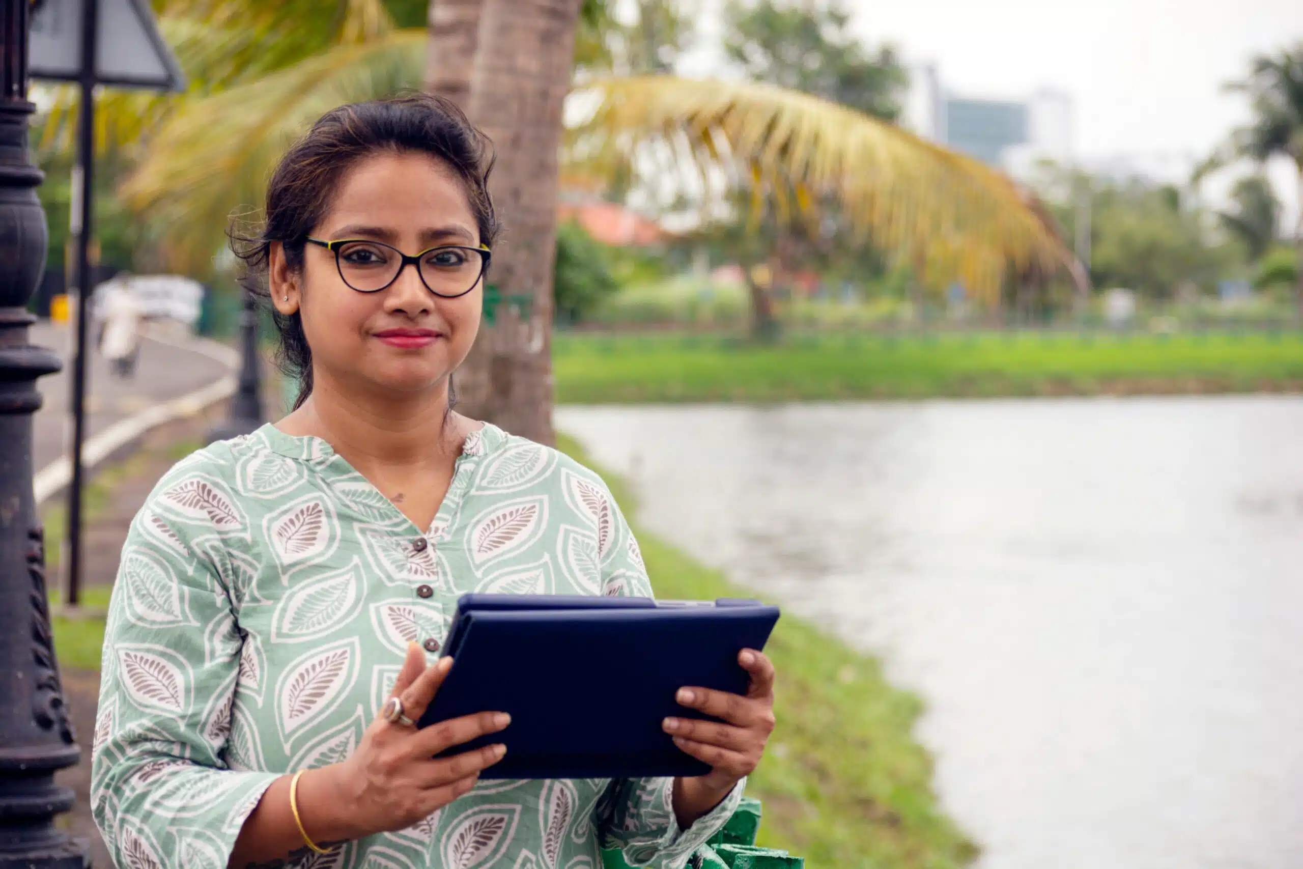 Woman looking into the camera, holding a tablet computer. With palm trees and a water body in the background.