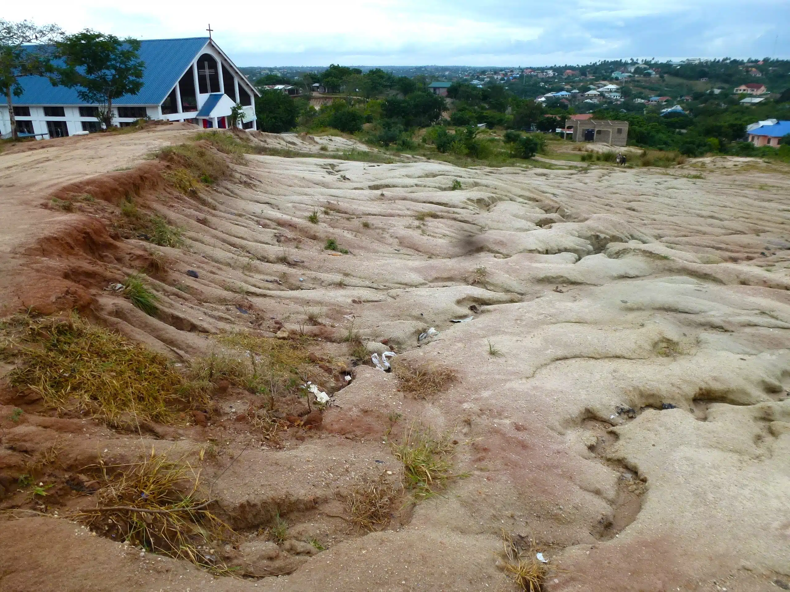 Reddish brown sediment in a dry, wide riverbed. Undulating surface. With church and trees in the background.