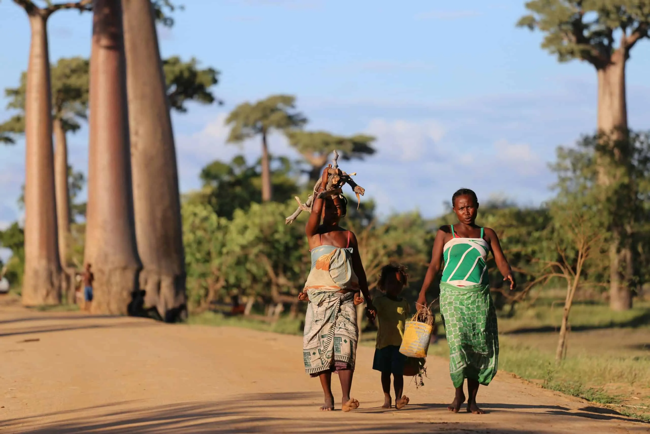 Women and child walking along dusty road. Large trees in the background and blue sky overhead.