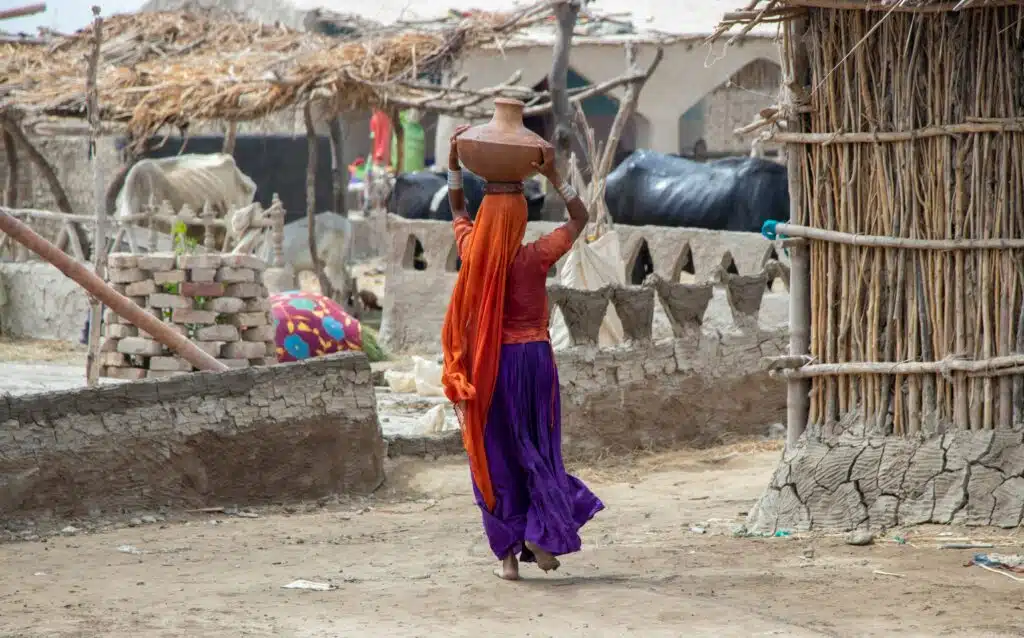Woman in orange and blue dress walking on brown sand, carrying matka (water vessel) on her head. Pakistan.