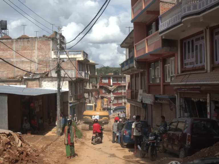 People walking and driving through earthquake damaged streets. Overhead wires hang above, damaged buildings on either side. Construction machinery can be seen in the background.