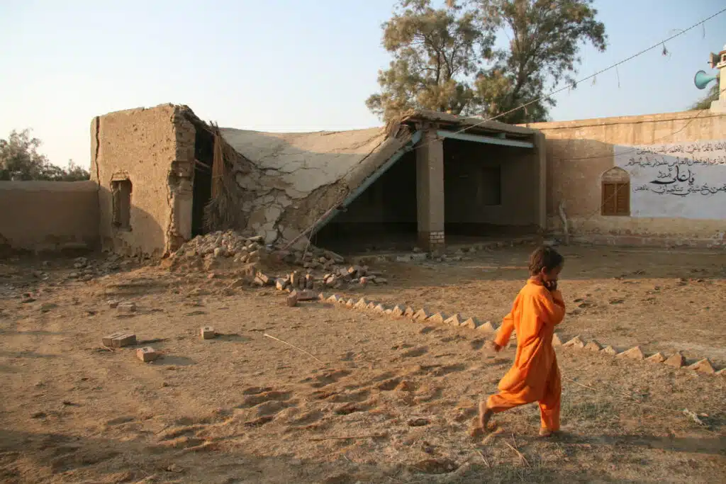 Young boy walking on dusty ground in front of flood damaged building, Pakistan