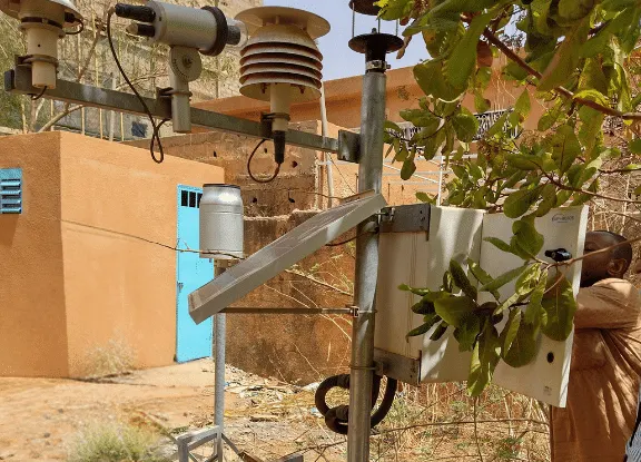 Person working on some hydromet equipment. Brightly coloured, orange building in the background. Foliage in the foreground