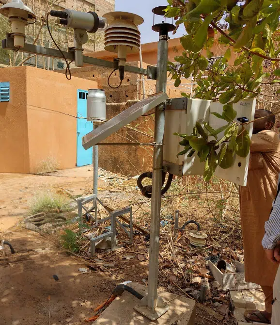 Person working on some hydromet equipment. Brightly coloured, orange building in the background. Foliage in the foreground