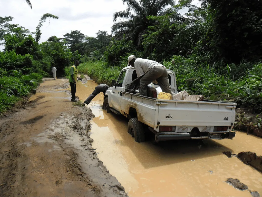 Pickup truck stuck in flooded, dirt road. Surrounded by lush tropical vegetation.