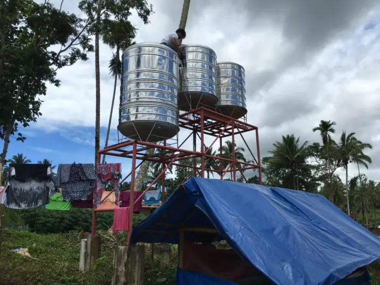 Water vessels providing temporary water supply to displacement camp. Temporary shelter in the foreground. Washing hanging to dry behind with palm trees lining the background.