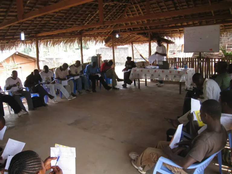 People seated in a circle in a large, straw roofed, open sided building. An instructor stands at the head of the circle with a whiteboard beside him.