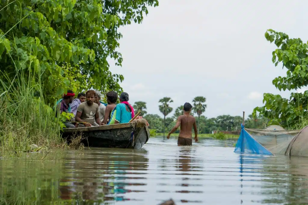Group of people, men and women on a boat travelling through flood water. A man is wading through the flood water beside them.