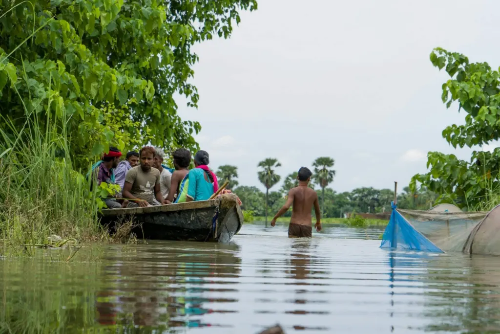 Group of people, men and women on a boat travelling through flood water. A man is wading through the flood water beside them.