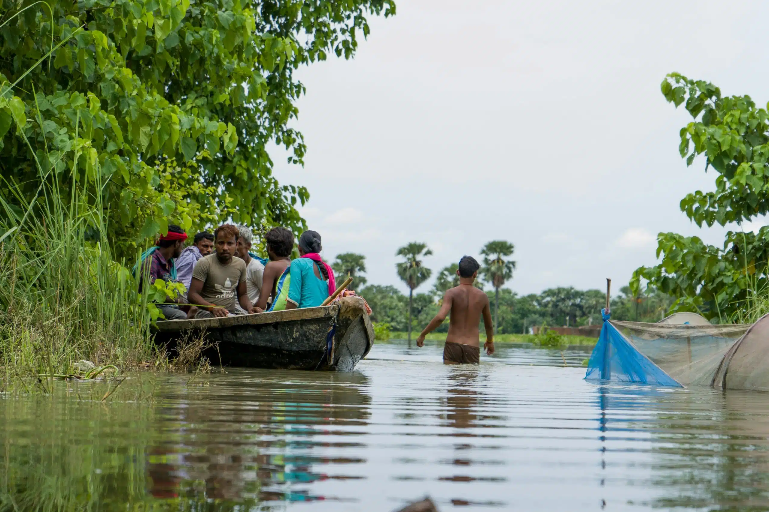 Group of people, men and women on a boat travelling through flood water. A man is wading through the flood water beside them.
