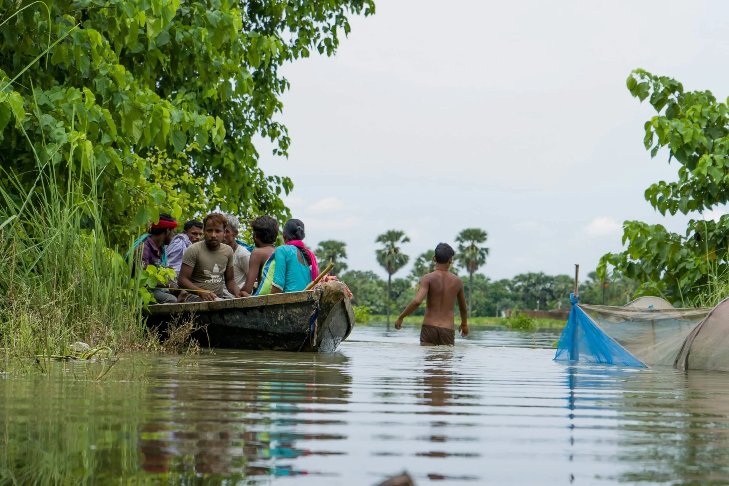 Group of people, men and women on a boat travelling through flood water. A man is wading through the flood water beside them.