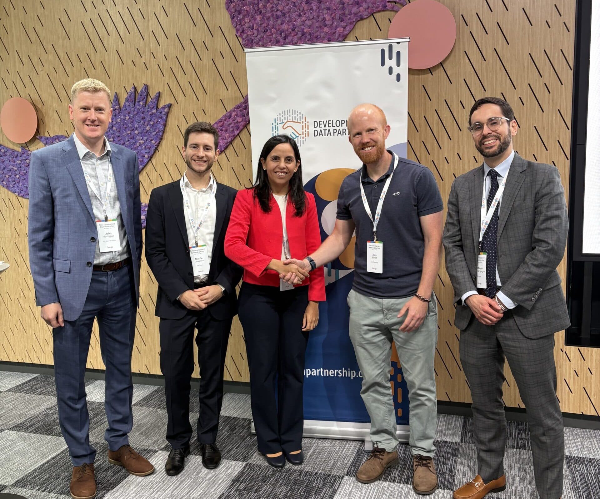 Alex Minett of JBA and Patricia Yañez-Pagans of IDB Invest shake hands in front of a Development Data Partnership banner at the Development Data Partnership Day event in New York. Three other men stand beside them, all smiling at the camera.