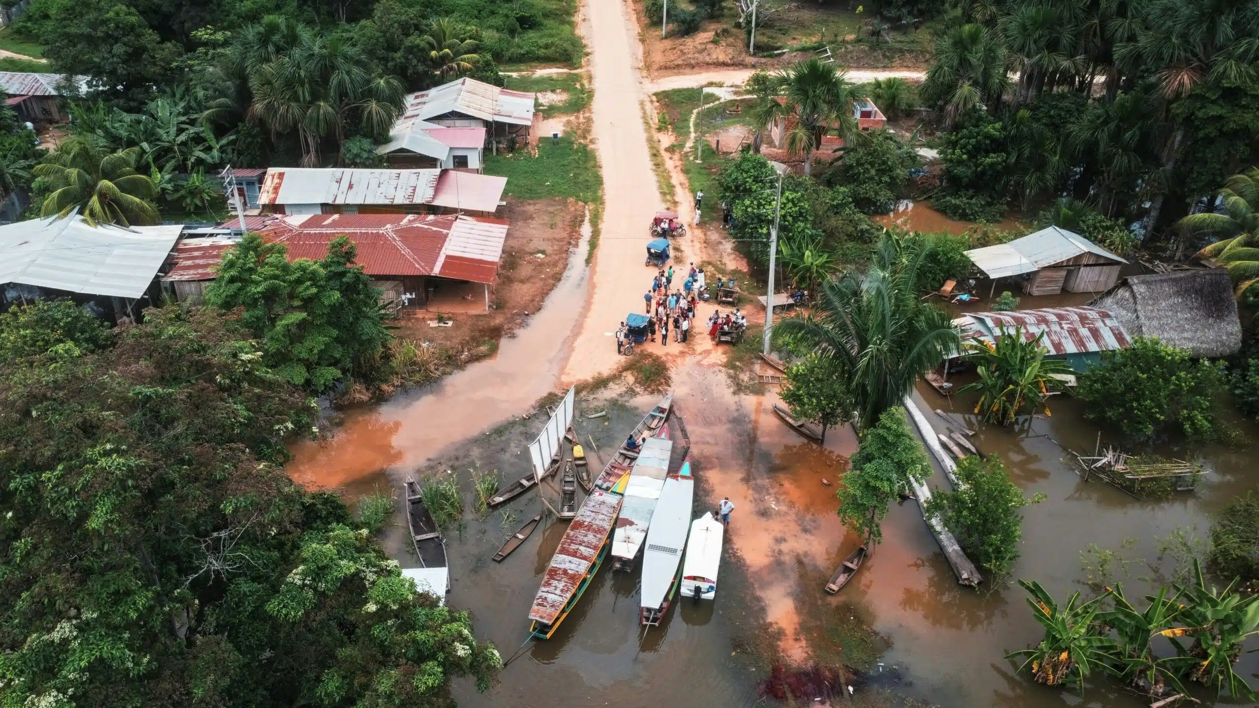 Arial photo of water inlet with a collection of boats. People are gathered at the junction of two, flooded, dirt roads. A cluster of houses with corrugated metal roofs are nestled in amongst trees and vegetation.