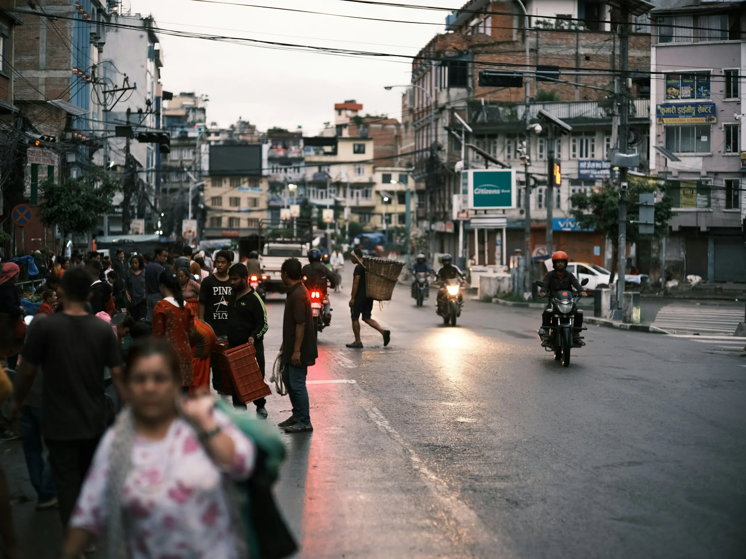 Bustling street scene in Nepal at dusk, with people walking, crossing the road, and scooters weaving past. Overhead wires stretch between buildings above the lively street.