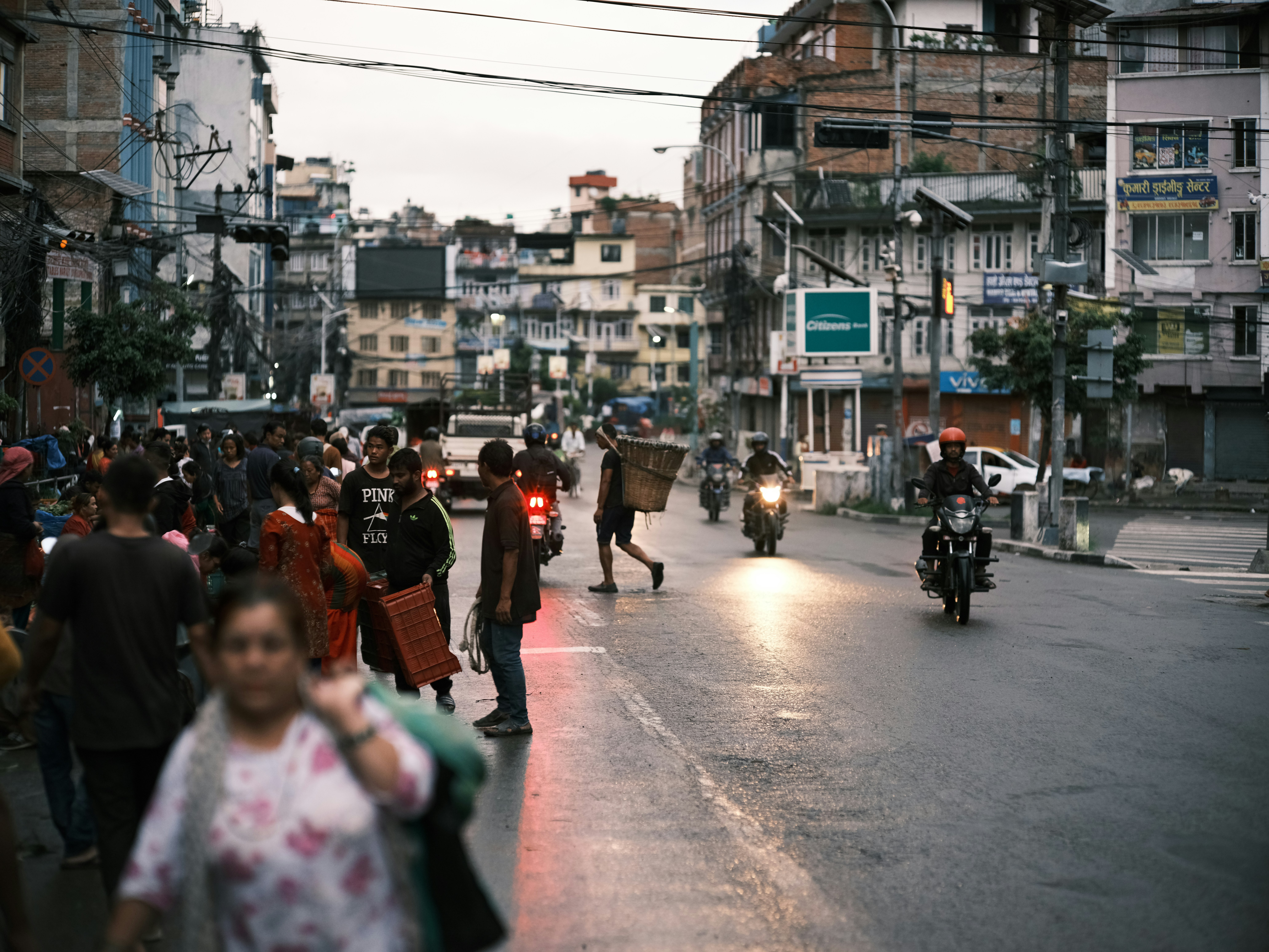 Bustling street scene in Nepal at dusk, with people walking, crossing the road, and scooters weaving past. Overhead wires stretch between buildings above the lively street.