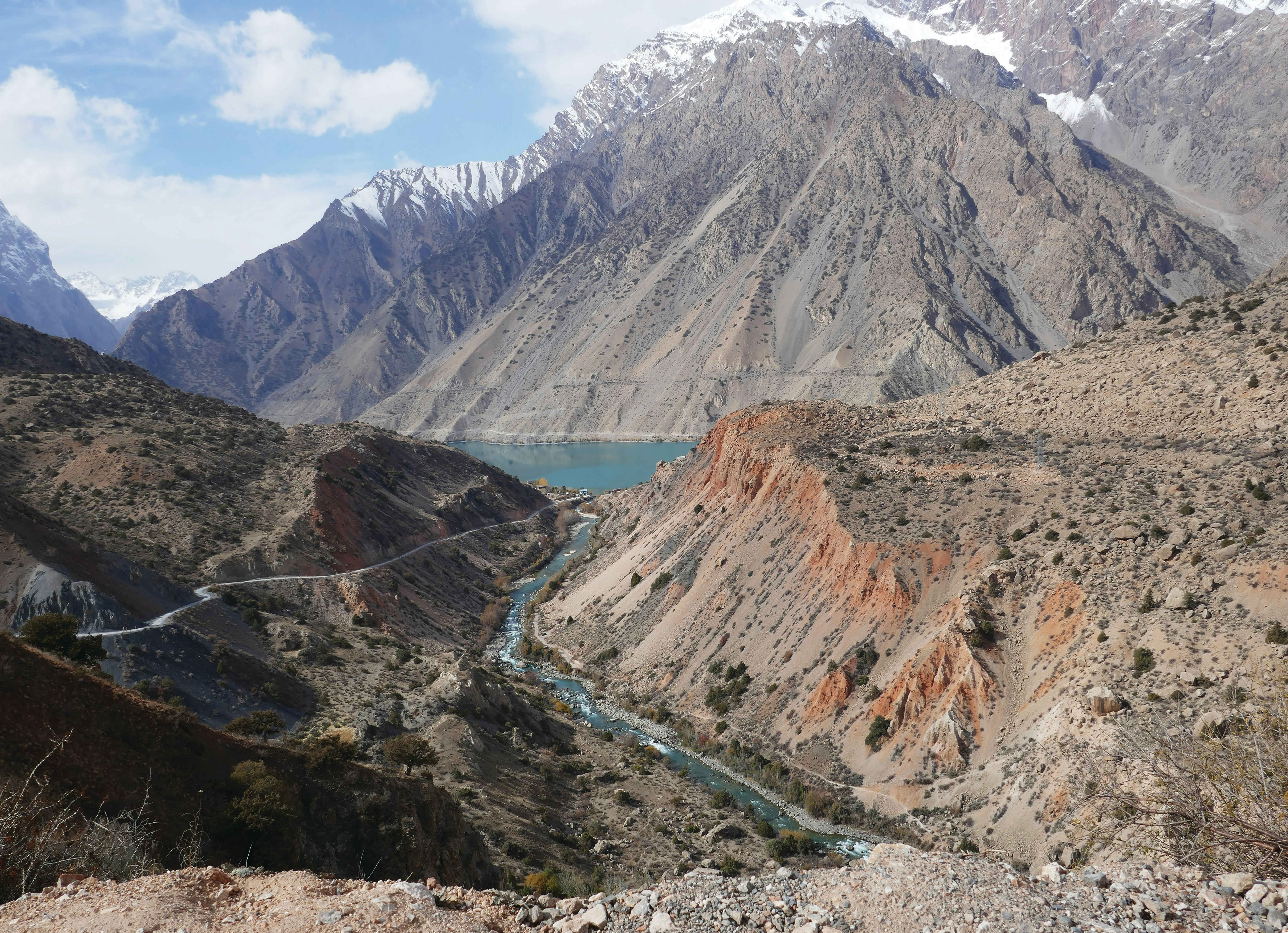 View from a mountain in Tajikistan looking down a dry, rugged valley with a river meandering through. A road follows the river along the lower reaches of the valley side. In the distance, a lake feeds the river, with snow-capped peaks rising beyond.