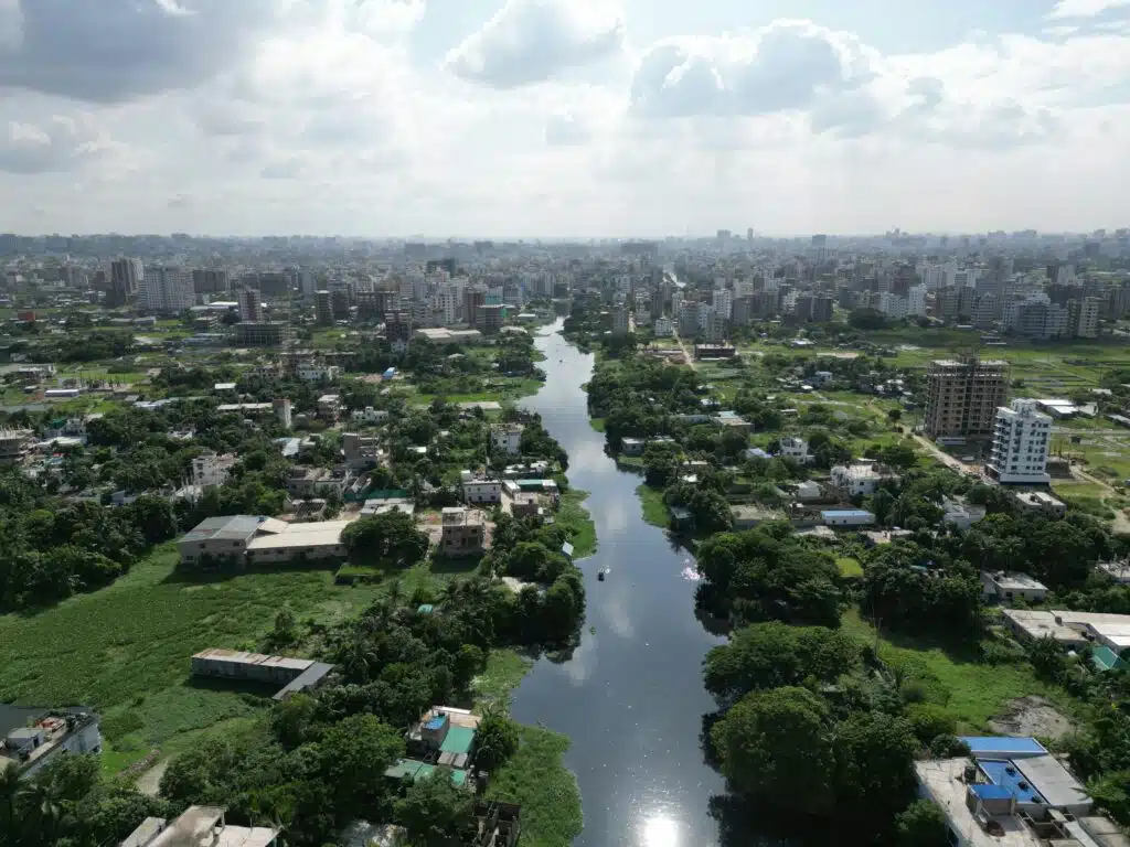 Aerial view of Dhaka, Bangladesh, showing a narrow river cutting through a densely populated urban area. The river is bordered by green vegetation and low-rise buildings in the foreground, with a dense skyline of high-rise buildings stretching into the horizon under a partly cloudy sky.