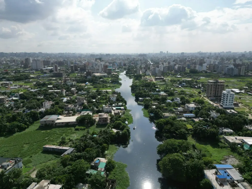 Aerial view of Dhaka, Bangladesh, showing a narrow river cutting through a densely populated urban area. The river is bordered by green vegetation and low-rise buildings in the foreground, with a dense skyline of high-rise buildings stretching into the horizon under a partly cloudy sky.