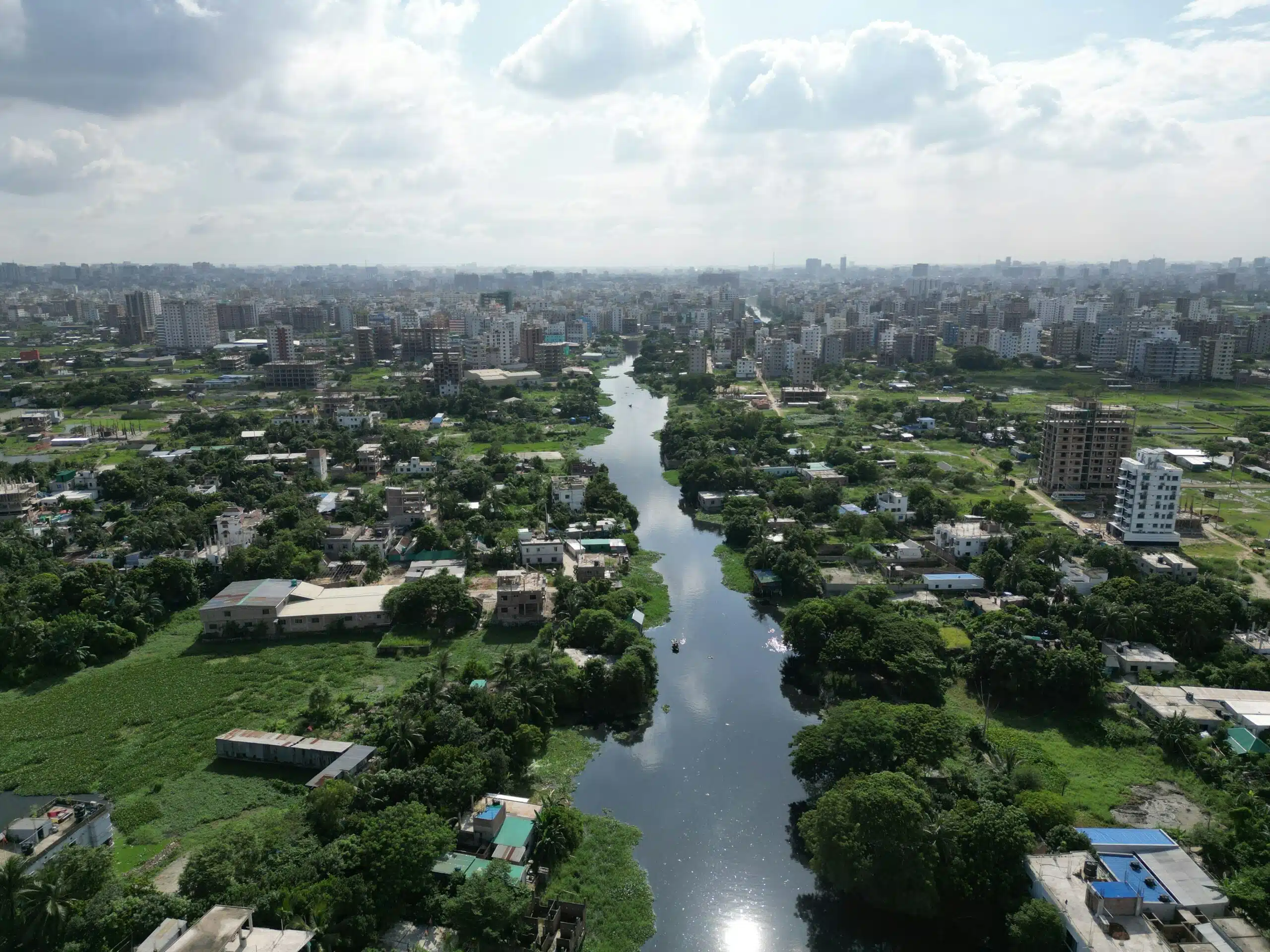 Aerial view of Dhaka, Bangladesh, showing a narrow river cutting through a densely populated urban area. The river is bordered by green vegetation and low-rise buildings in the foreground, with a dense skyline of high-rise buildings stretching into the horizon under a partly cloudy sky.