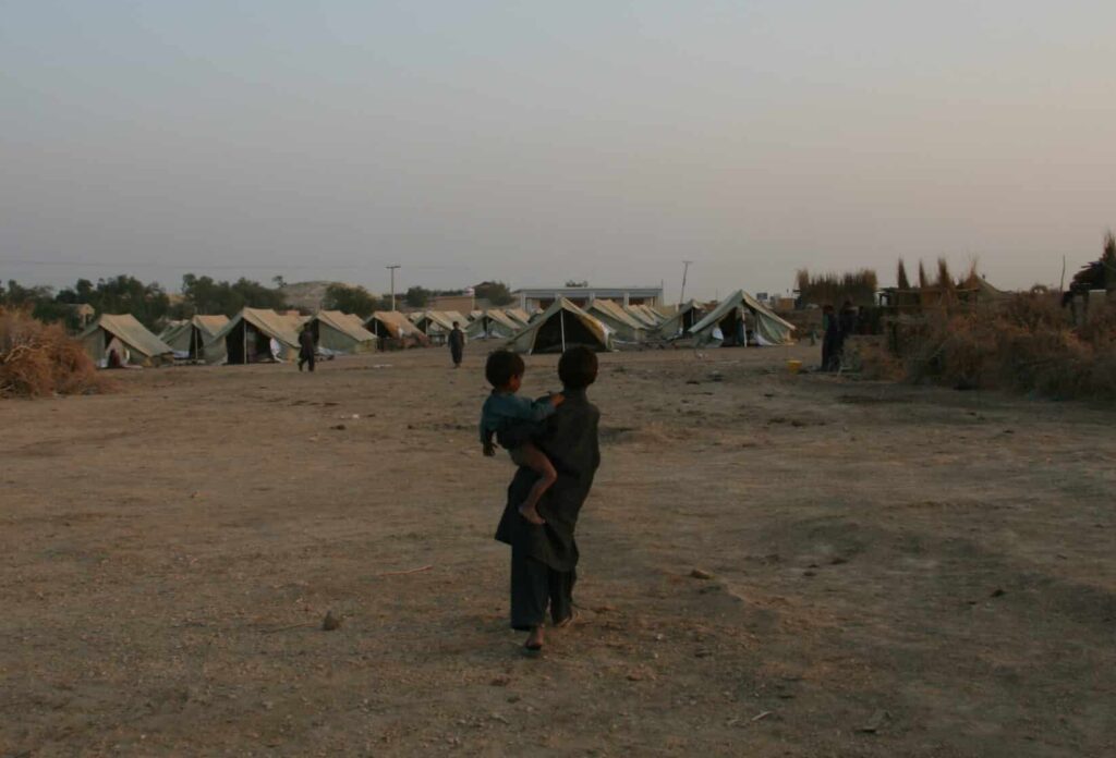 Two children in a dry, dusty landscape walking towards a temporary tent settlement, with makeshift shelters and sparse vegetation under a clear evening sky.