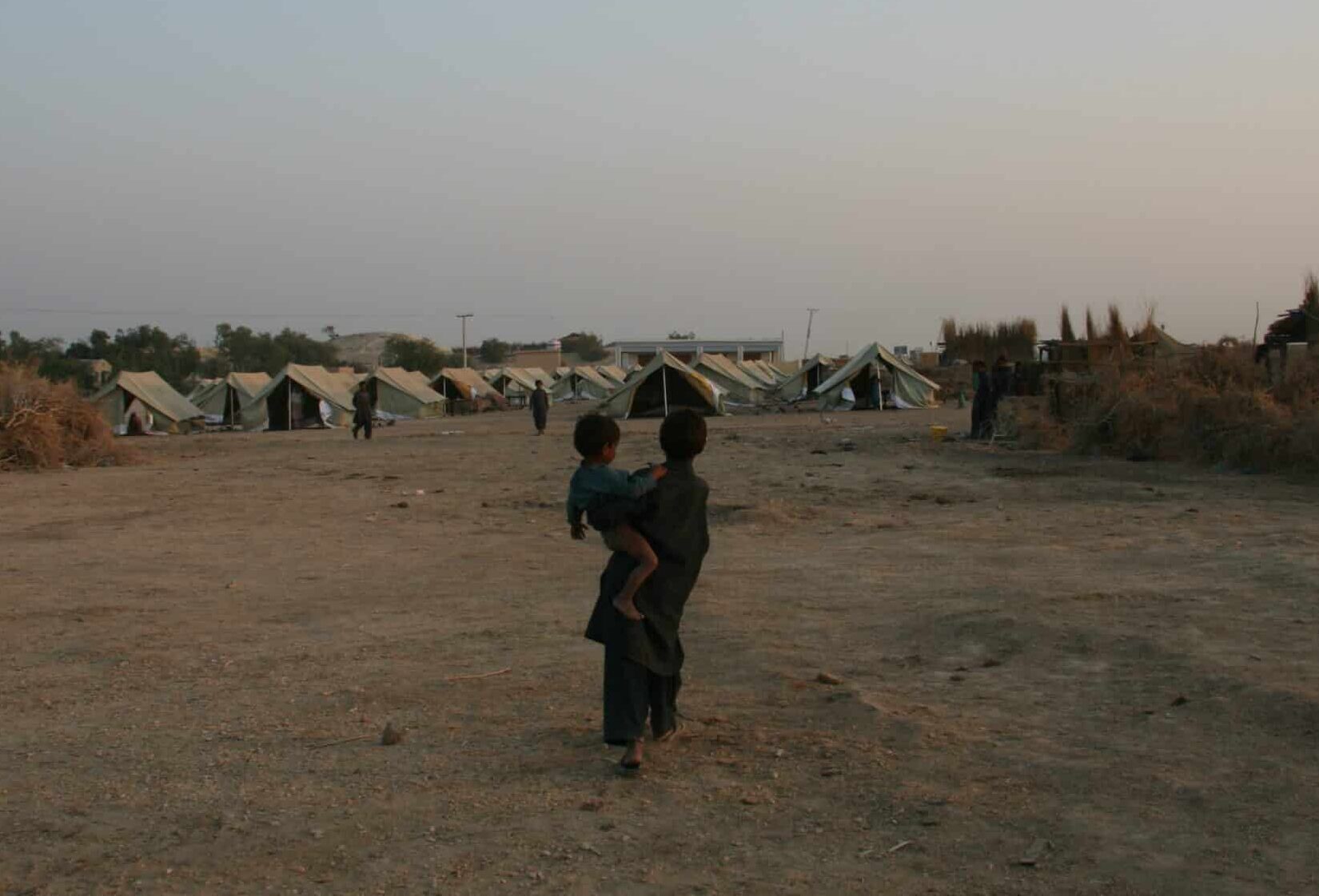 Two children in a dry, dusty landscape walking towards a temporary tent settlement, with makeshift shelters and sparse vegetation under a clear evening sky.