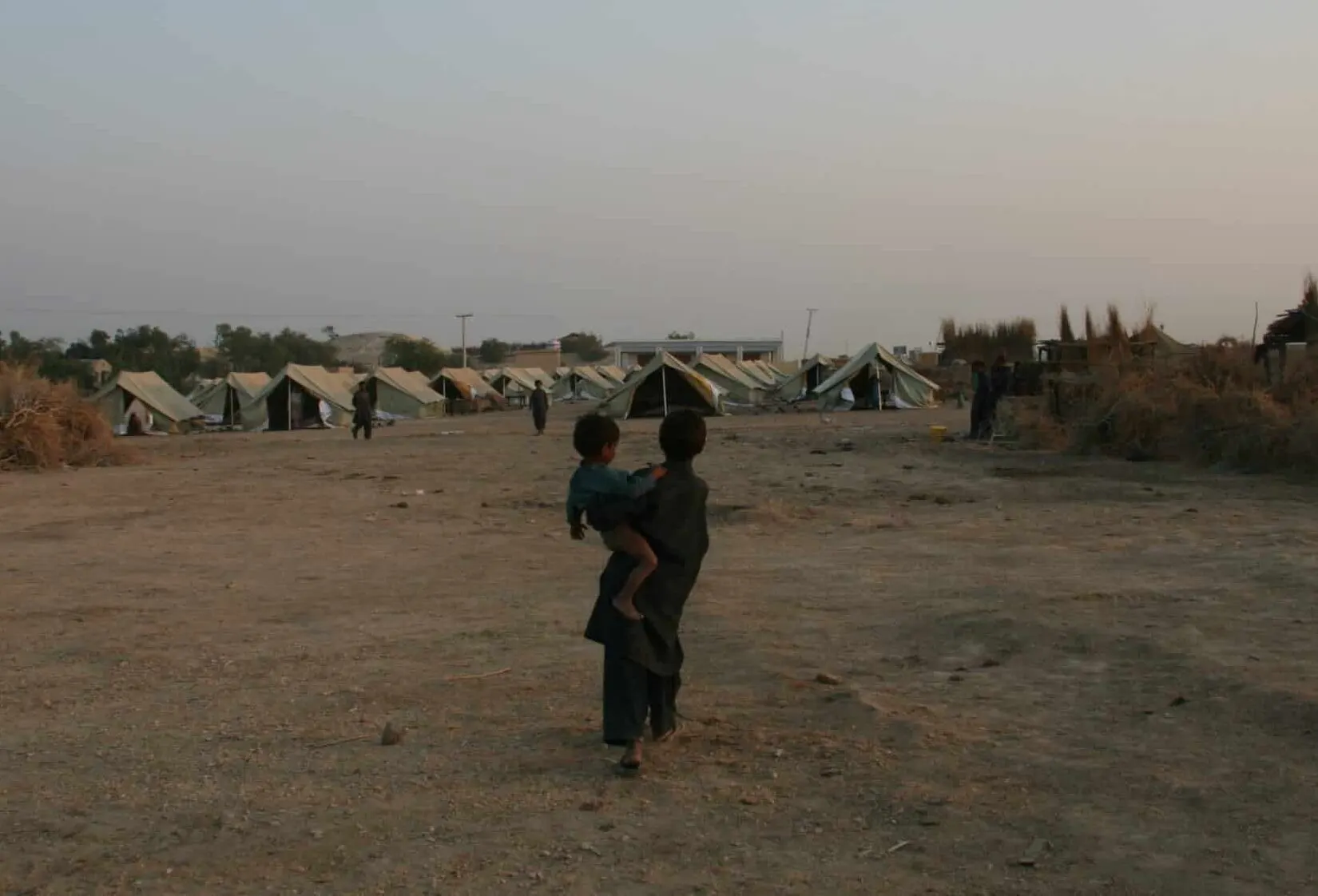 Two children in a dry, dusty landscape walking towards a temporary tent settlement, with makeshift shelters and sparse vegetation under a clear evening sky.