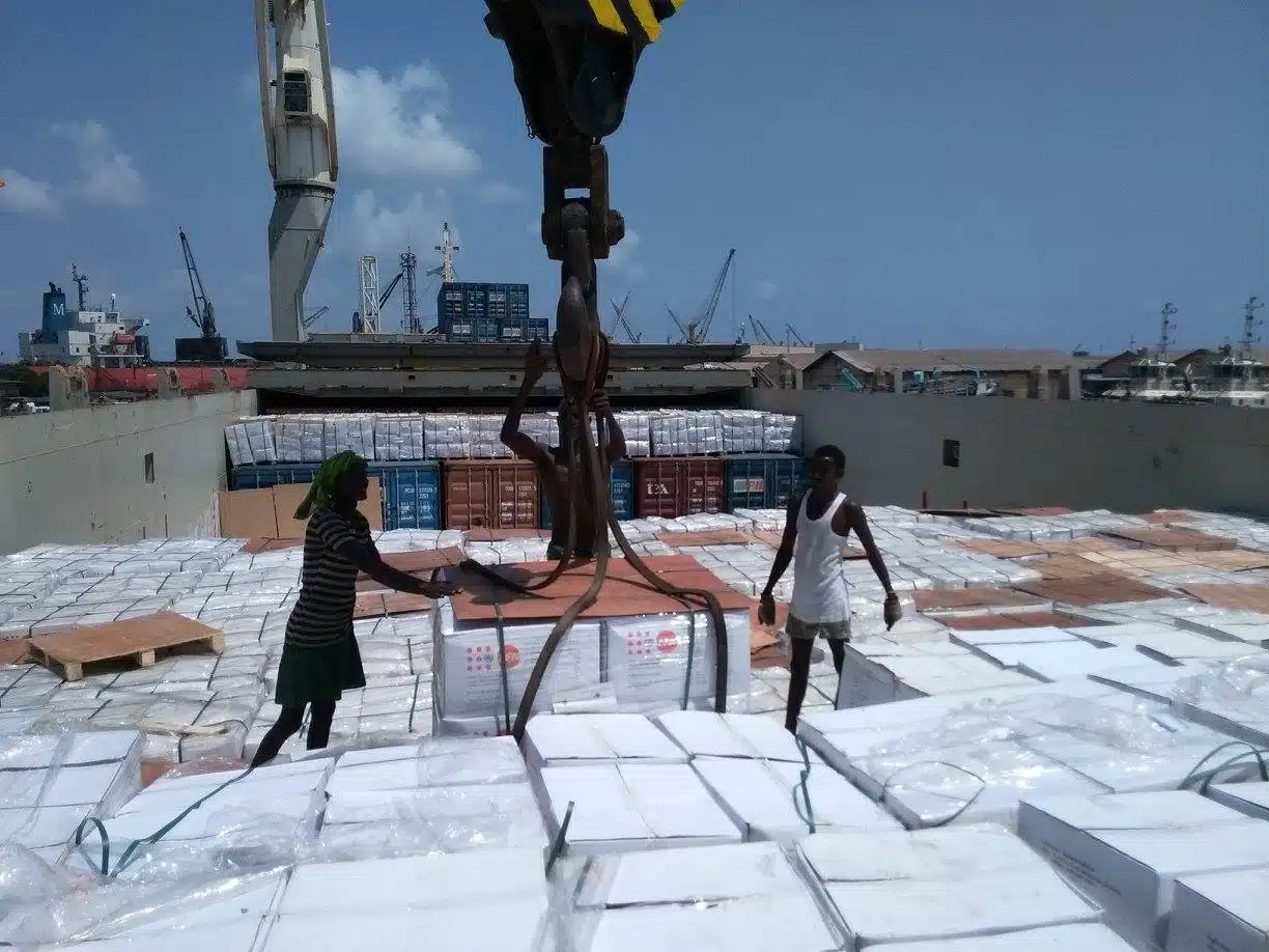 Workers on a cargo ship preparing pallets of boxed humanitarian supplies for unloading at a busy port, with cranes and shipping containers in the background.