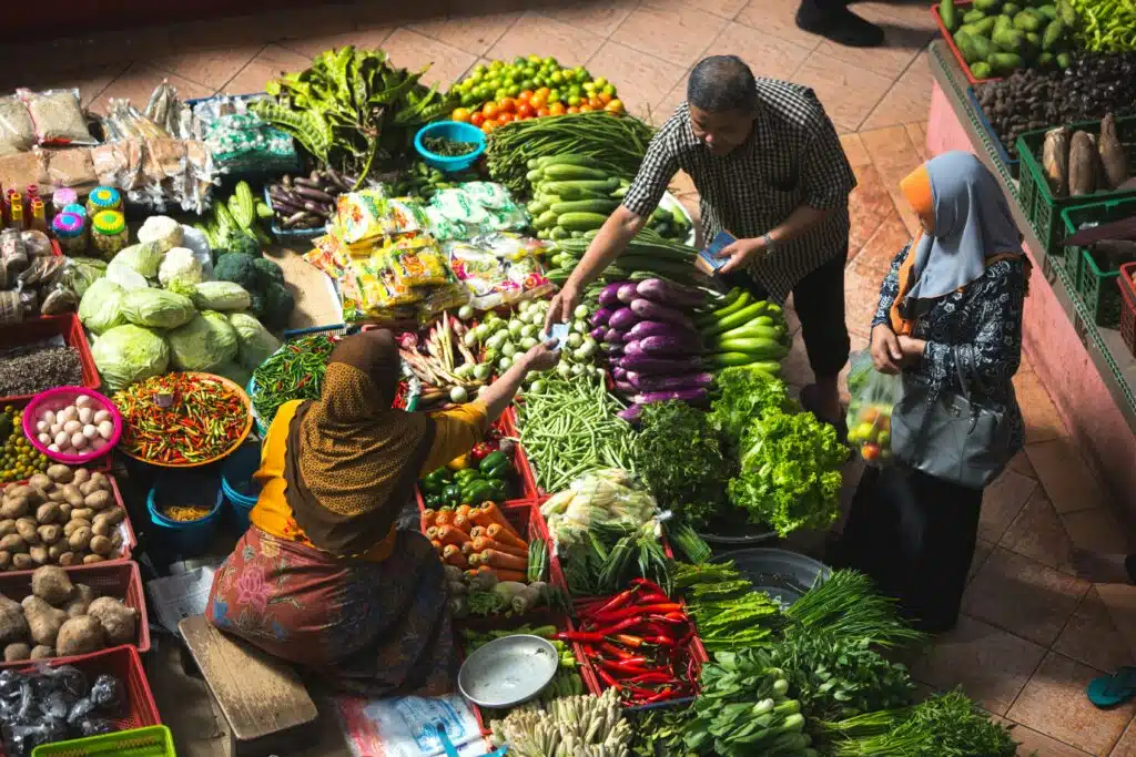 Vibrant market scene with a female vendor selling fresh vegetables to a customer, surrounded by colourful produce including chillies and leafy greens.