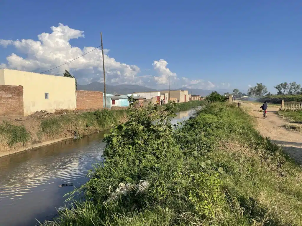 A small canal runs alongside a dirt path and houses with brick and painted walls in a rural community. Green vegetation grows along the canal, and a child rides a bicycle down the path under a bright blue sky with scattered clouds.
