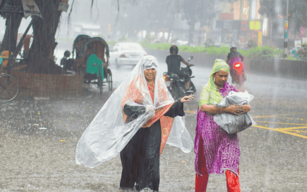 Two women walking through heavy rain and street flooding, using plastic coverings to shield themselves, with rickshaws and traffic in the background.