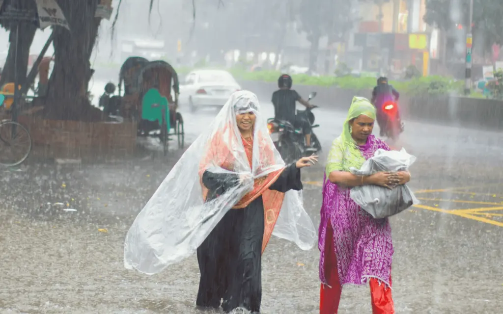 Two women walking through heavy rain and street flooding, using plastic coverings to shield themselves, with rickshaws and traffic in the background.