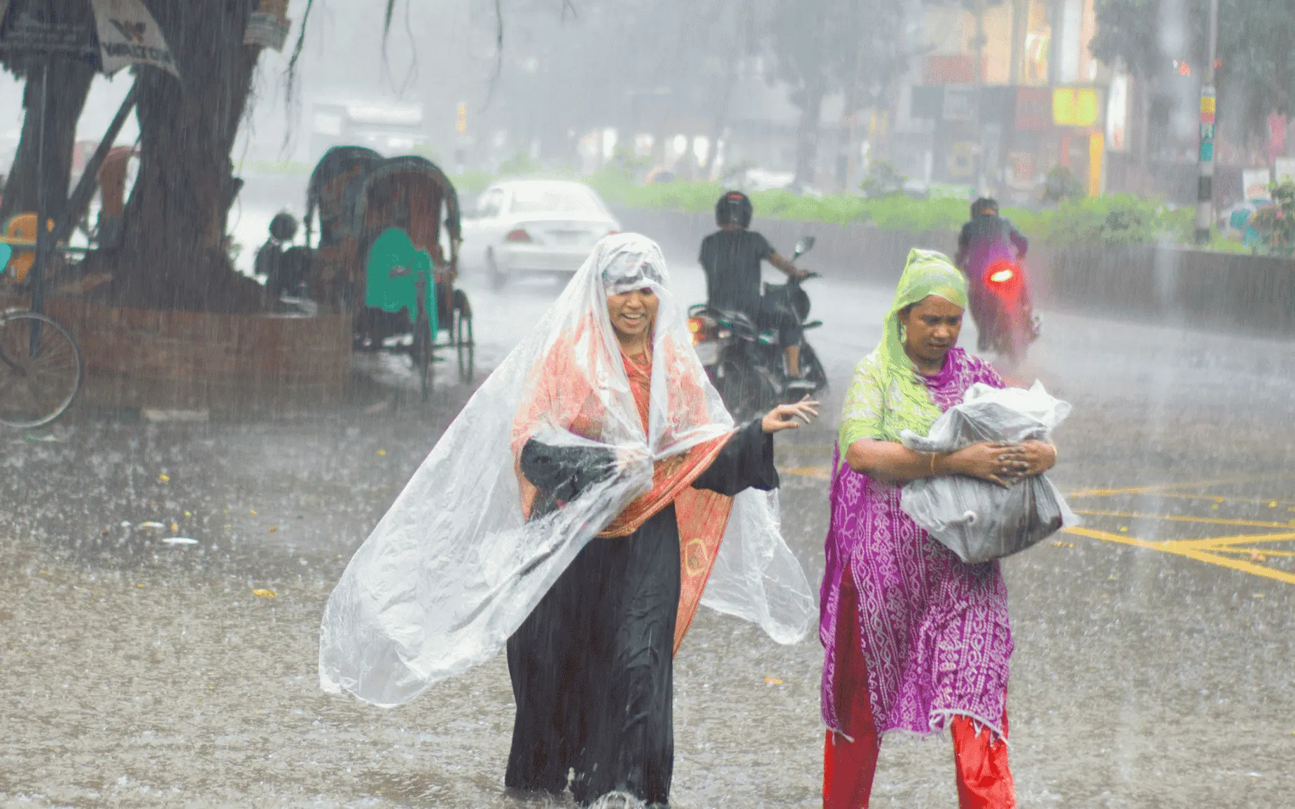 Two women walking through heavy rain and street flooding, using plastic coverings to shield themselves, with rickshaws and traffic in the background.