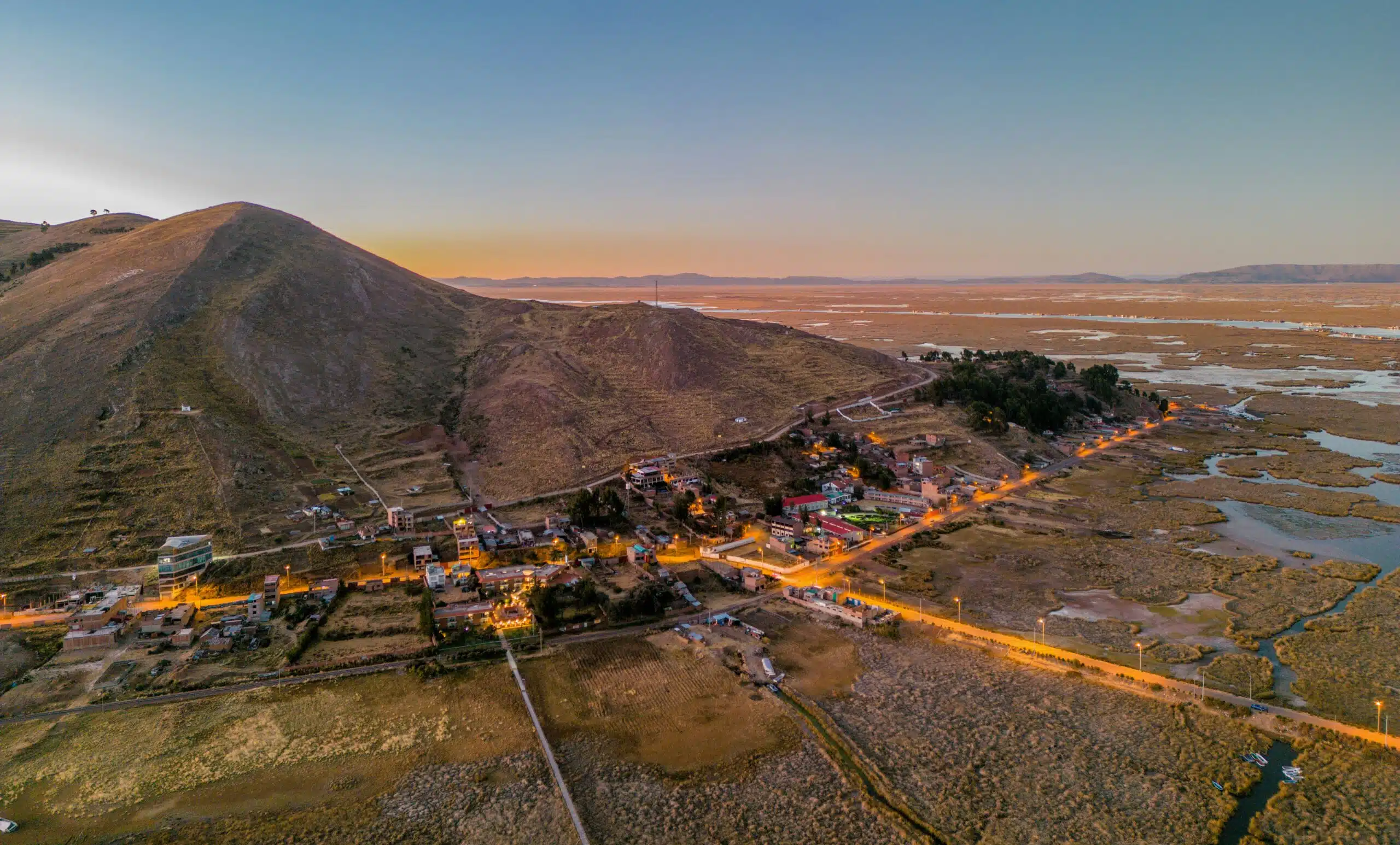 Aerial view of a rural town and surrounding hills at sunset near Lake Titicaca, Peru, showing roads, buildings, and wetland areas under soft golden light.