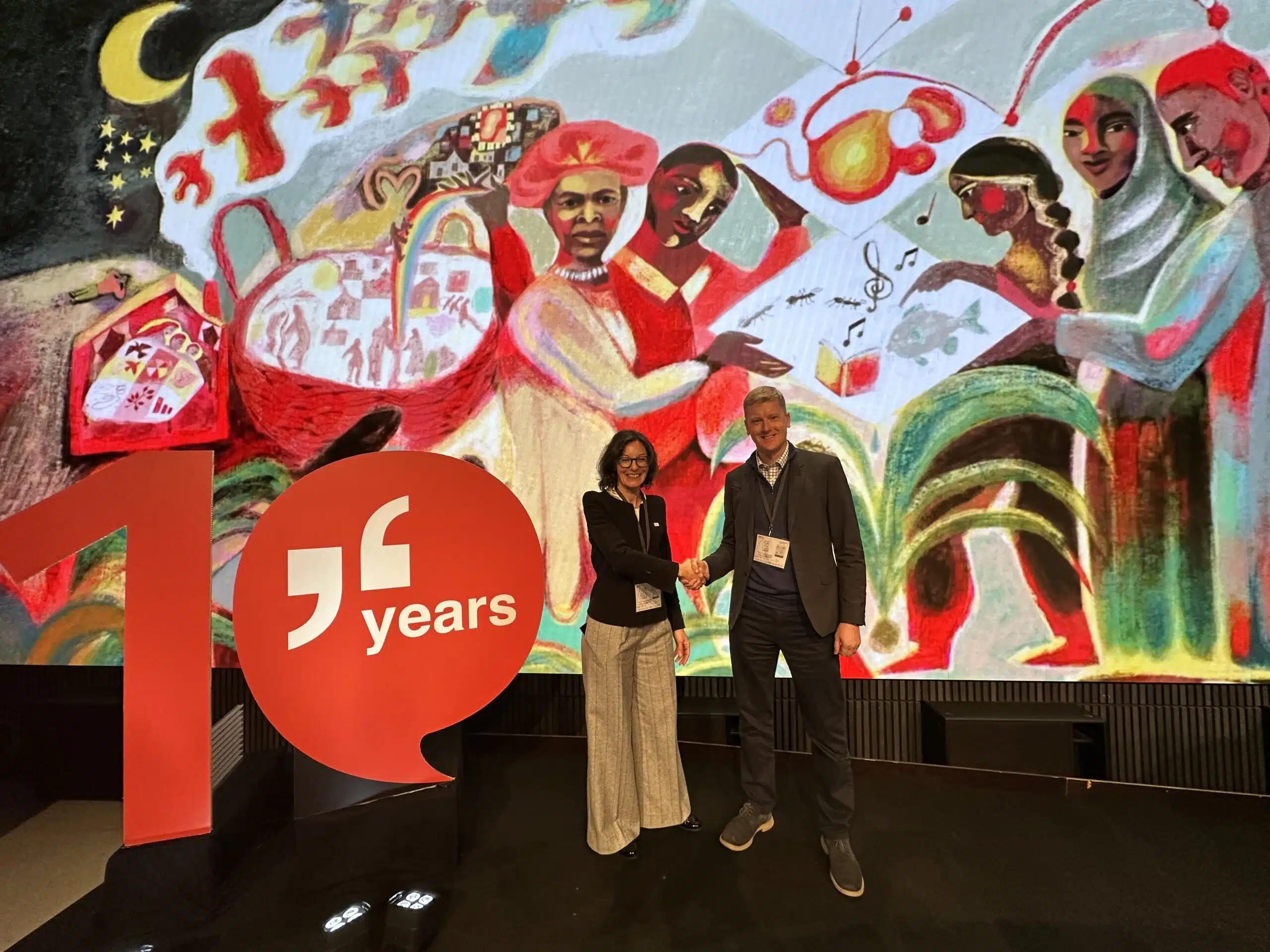 Alessandra Gilotta of the Anticipation Hub and Joh Bevington of JBA Global Resilience shake hands at the 13th Global Dialogue Platform of Anticipatory Humanitarian Action, standing in front of a large illustrated backdrop and a red “10 years” installation.