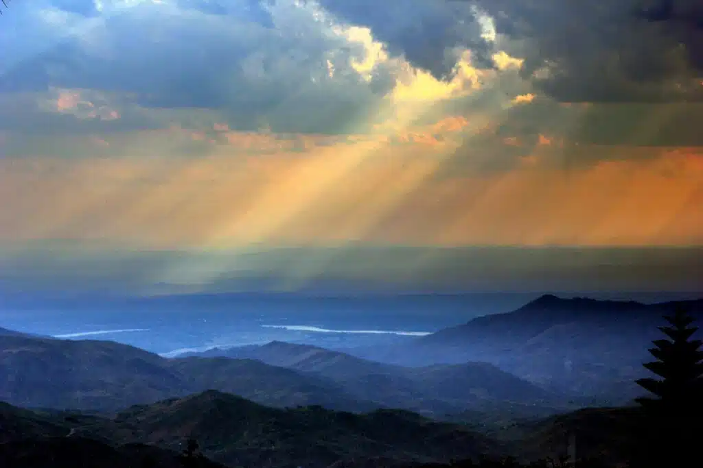 Sunlight streaming through dramatic clouds over layered mountain ranges and a distant river at sunset.