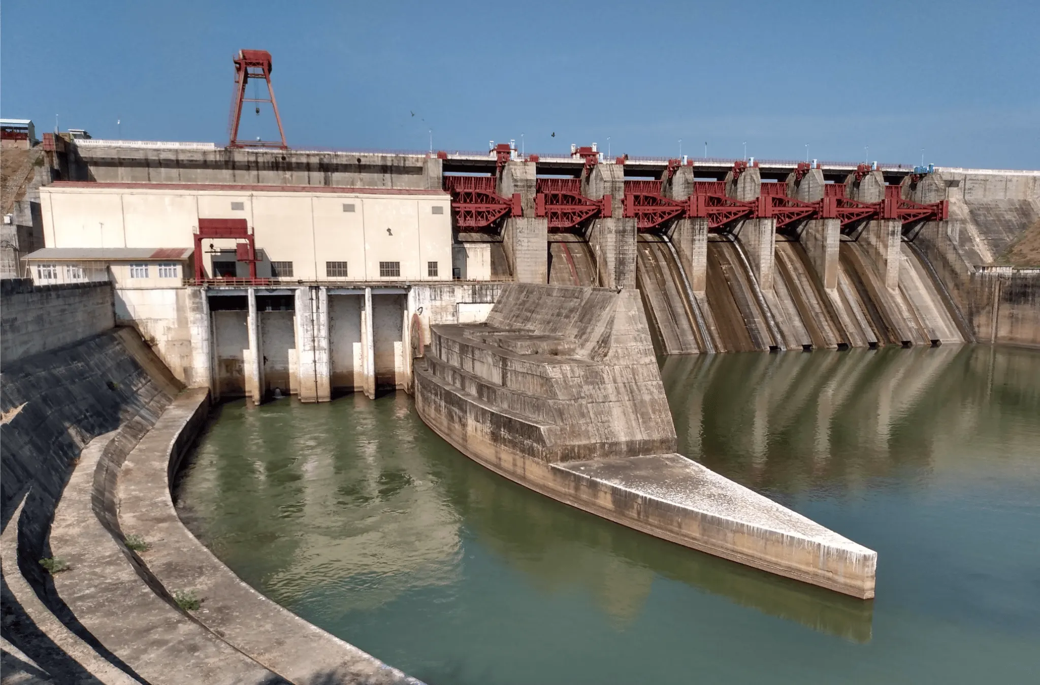 View of concrete dam in Myanmar from a downstream perspective. The sky is blue, the water brown, the dam is cream and red.