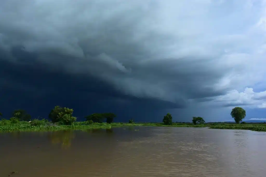 Large, dark thunderstorm clouds sweeping across the sky above a calm river, with trees and vegetation along the distant shoreline.