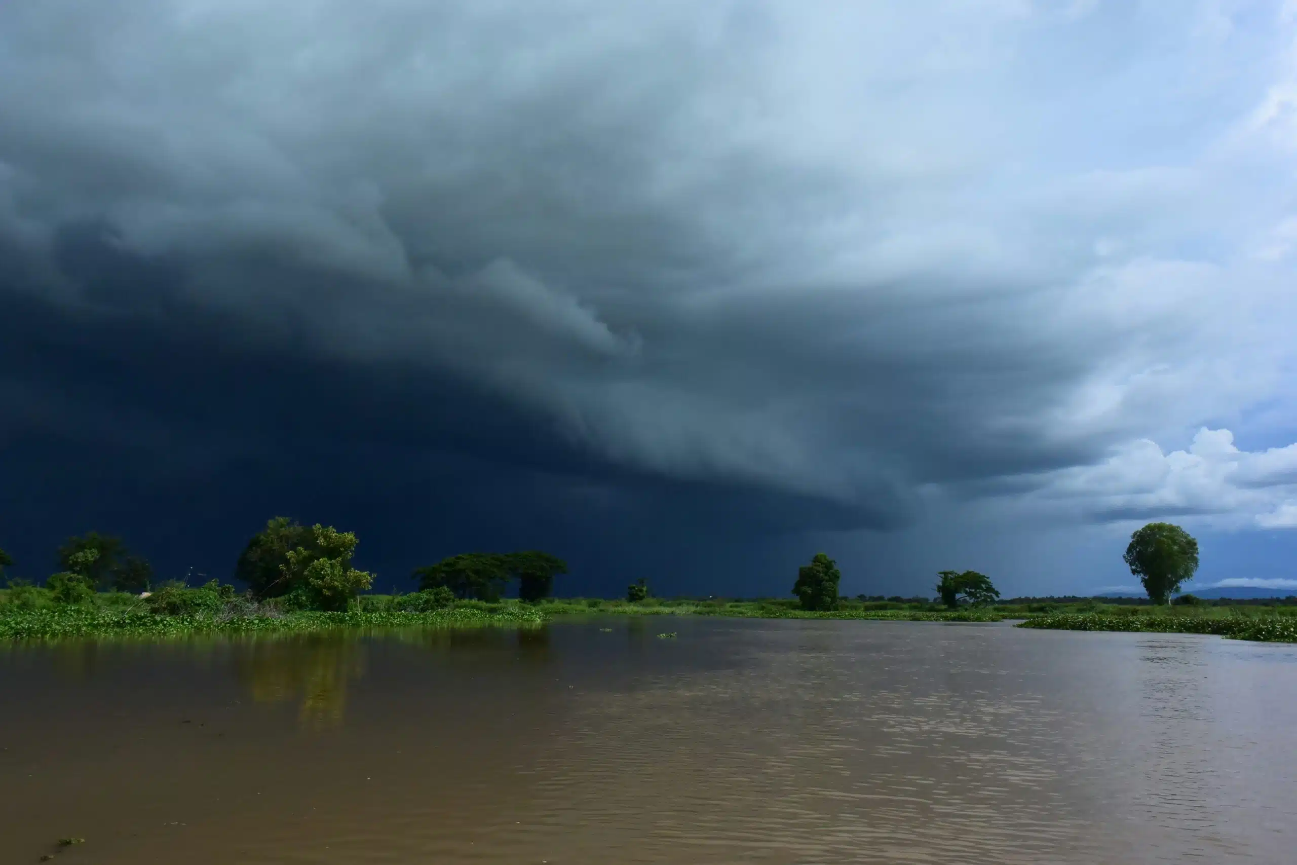 Large, dark thunderstorm clouds sweeping across the sky above a calm river, with trees and vegetation along the distant shoreline.