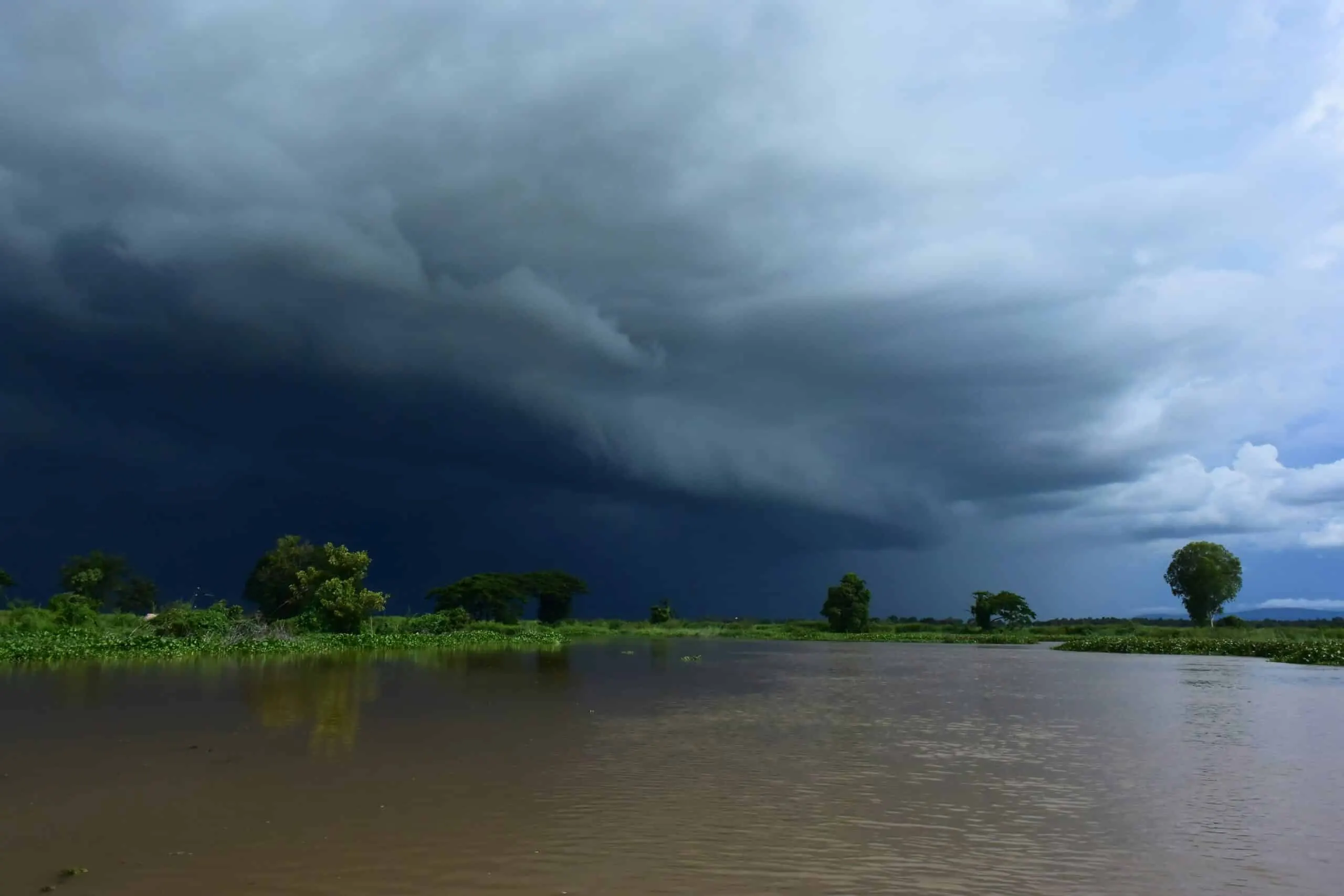 Large, dark thunderstorm clouds sweeping across the sky above a calm river, with trees and vegetation along the distant shoreline.