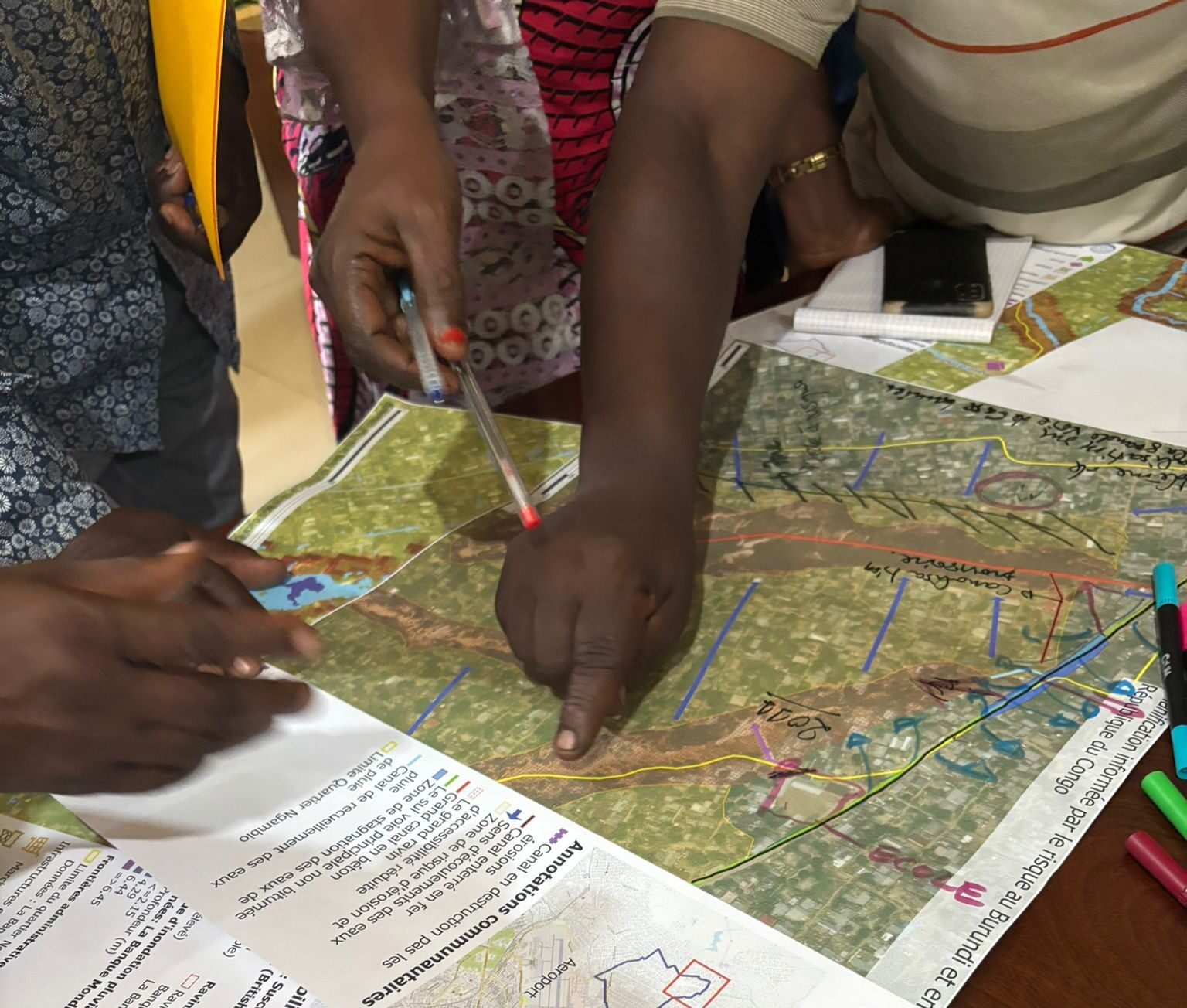 Hands of several participants pointing at and marking a large printed map during a community mapping activity, with pens and documents spread across the table.