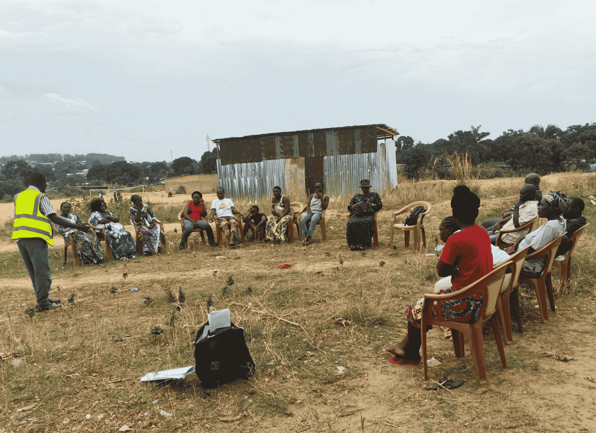 A community meeting taking place outdoors, with a group of people seated in a wide circle on plastic chairs in a dry, open field, while a facilitator in a high‑visibility vest speaks to them. A small corrugated metal building stands in the background.