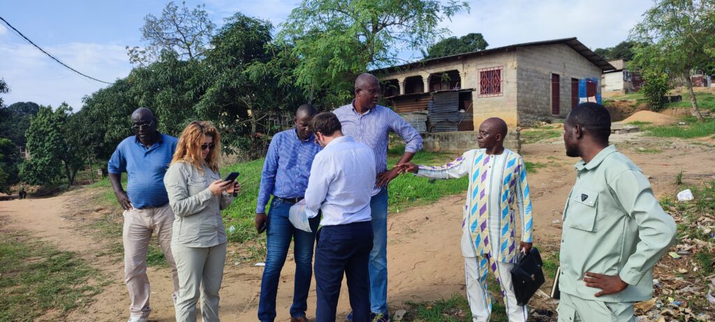A group of people standing together outdoors on a dirt path, discussing something while looking at documents or a device, with trees and simple buildings in the background.