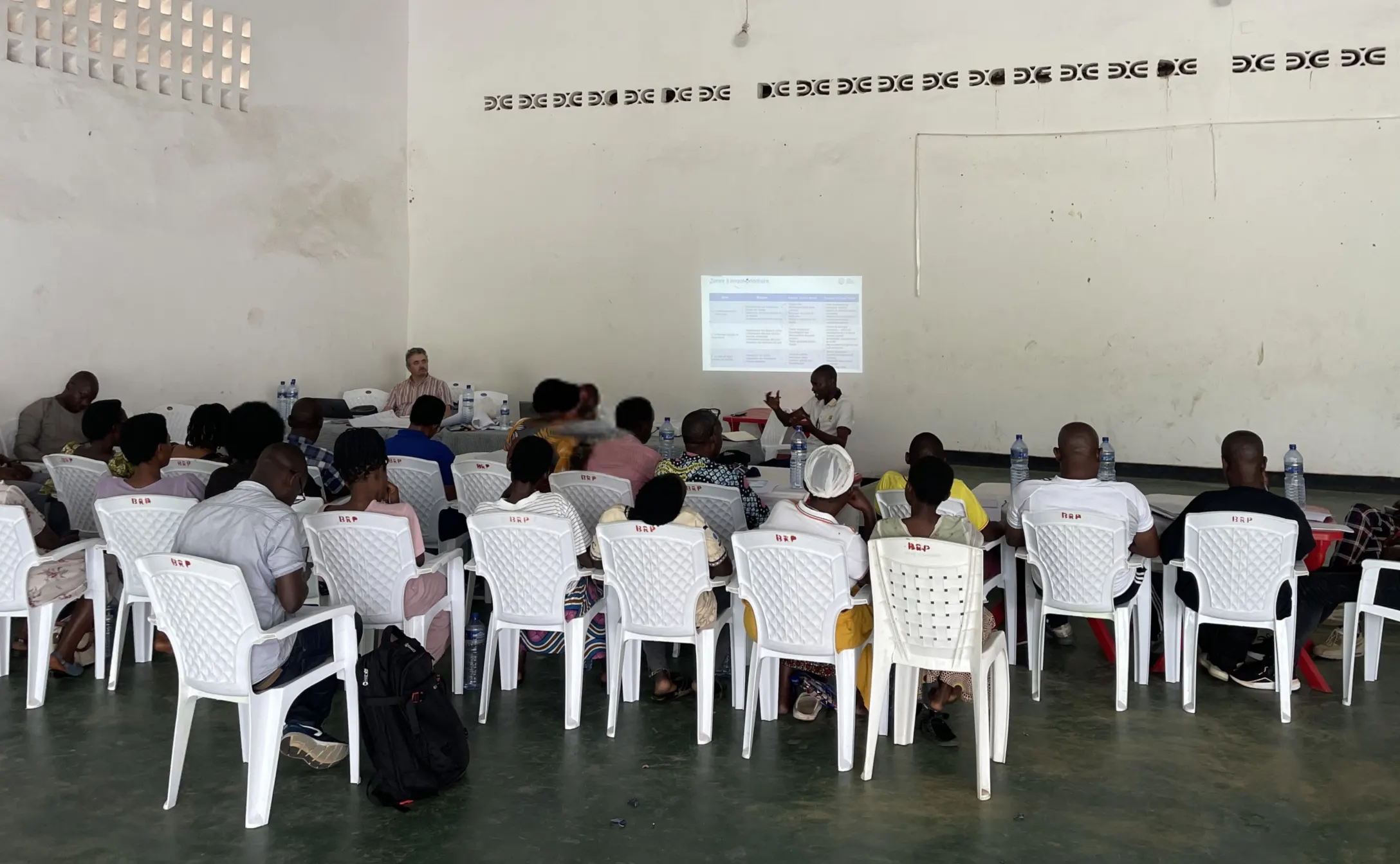 Participants seated on white chairs inside a large hall, attending a workshop with a presentation projected onto the wall at the front of the room.
