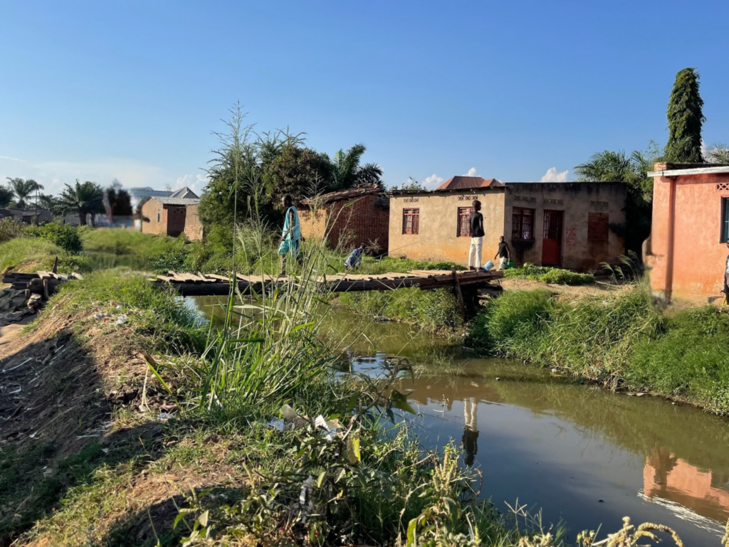 People walking and standing near a small water channel in a residential area, with houses and vegetation alongside the channel.