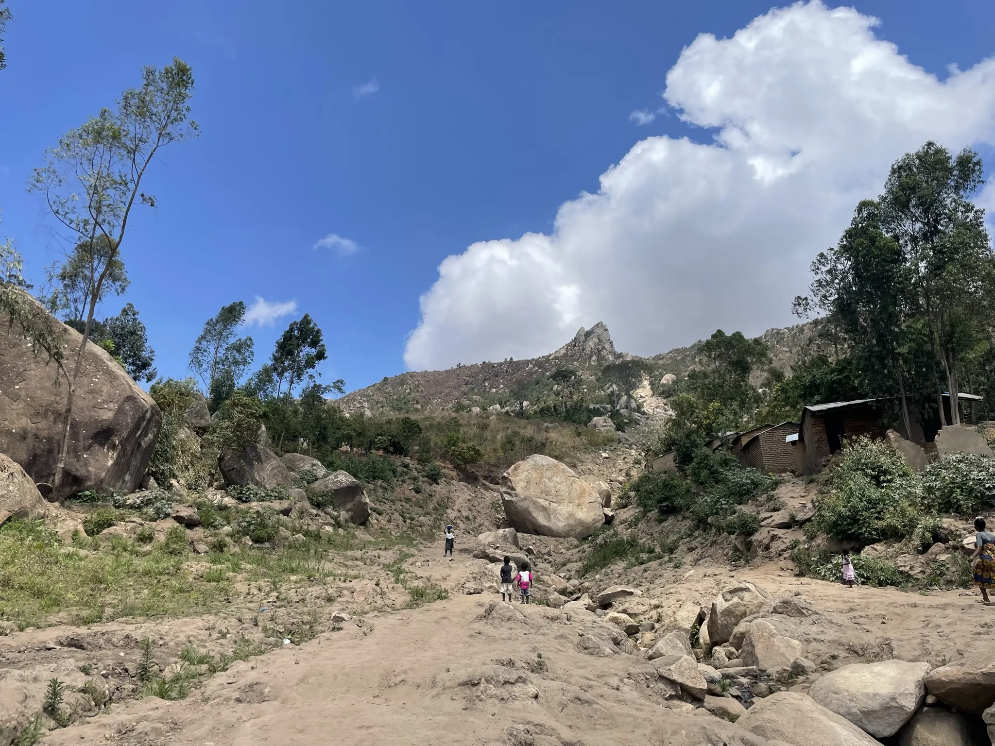 Rocky channel pathway through a hillside landscape, with scattered boulders, trees, and people walking.