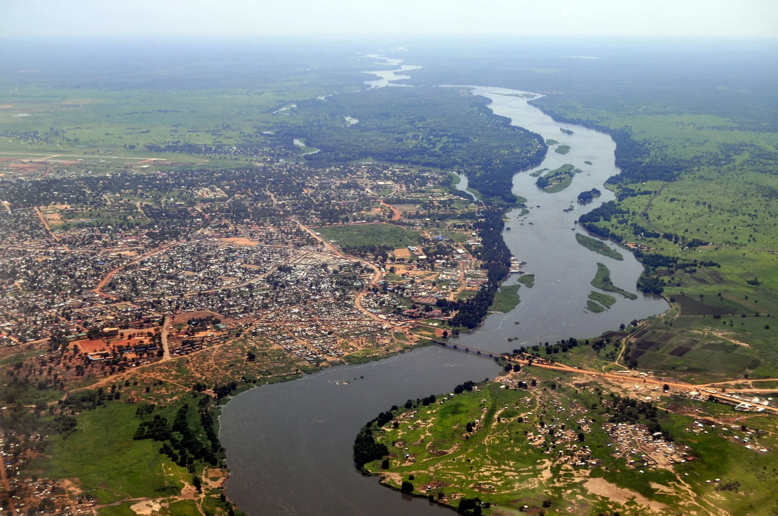 Aerial view of an urban settlement (Juba, South Sudan) beside a branching river system, showing dense buildings, surrounding fields, and exposed floodplain areas.