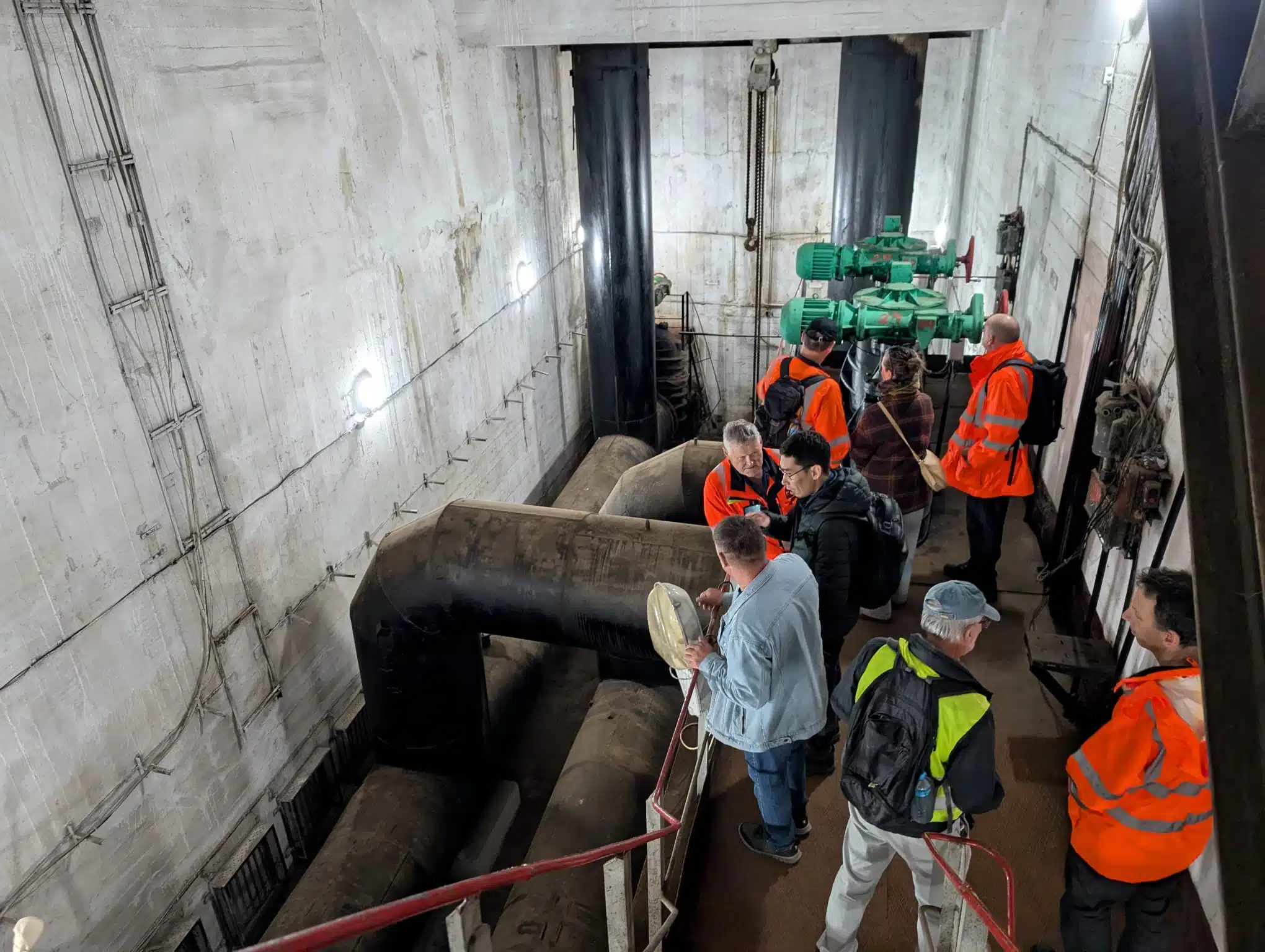 Group of engineers inspecting large pipes and control equipment inside a dam or water infrastructure facility, viewed from a walkway above the machinery.