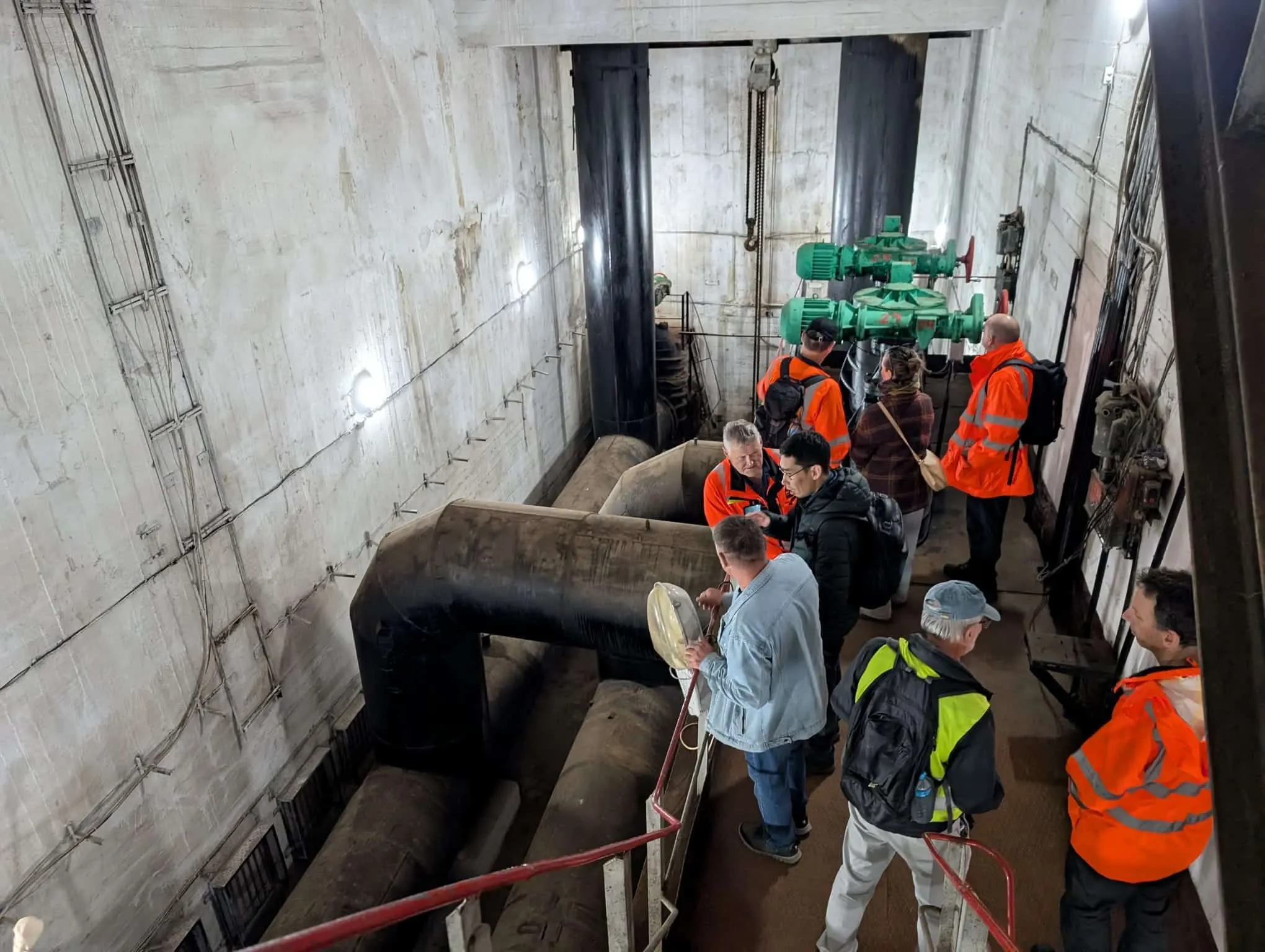 Group of engineers inspecting large pipes and control equipment inside a dam or water infrastructure facility, viewed from a walkway above the machinery.